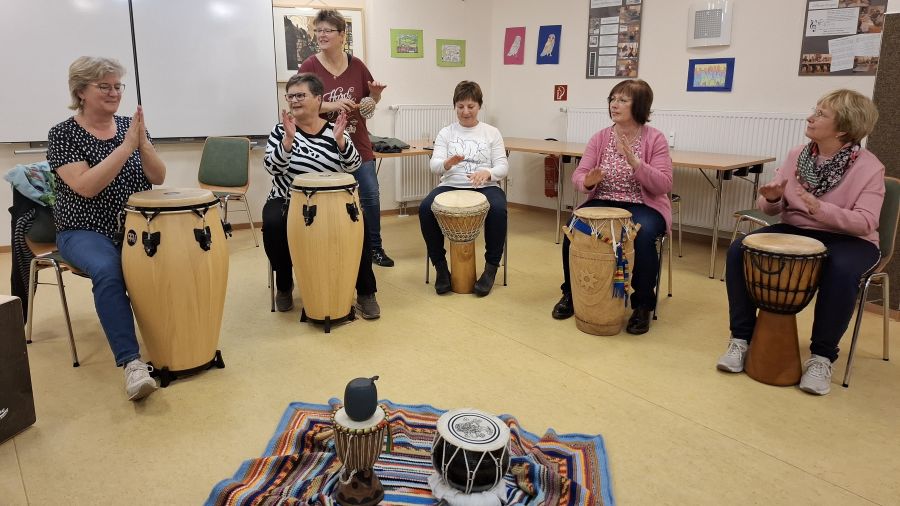 Das Foto zeigt Frauen beim Trommel-Workshop, wie sie mit den Händen klatschen und auf vor ihnen stehende Trommeln schlagen. 